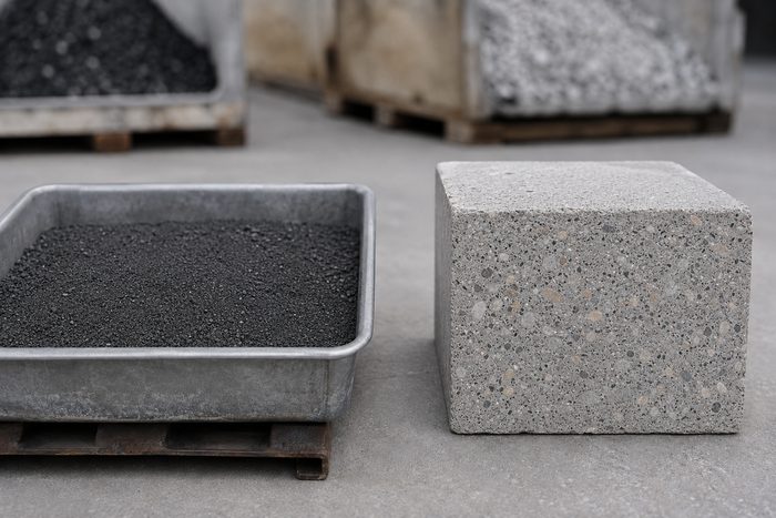 A bin of dark granular reclaimed-carbon feedstock beside a finished cured concrete block flecked with dark aggregate